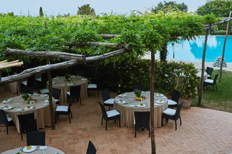 An elegant outdoor dining setup at an Amalfi Coast villa, featuring round tables set with lemons under a vine-covered pergola beside a blue swimming pool.