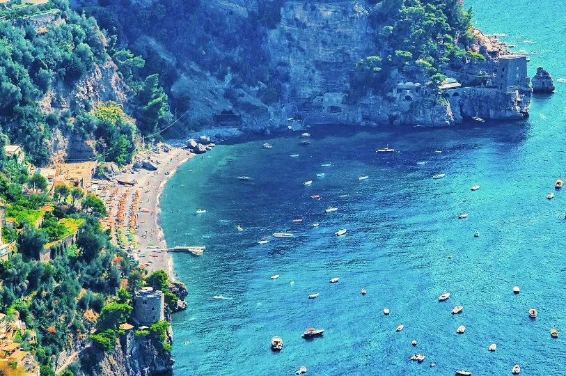 A high-angle view of a picturesque cove on the Amalfi Coast, showing a secluded beach lined with orange umbrellas, a historic stone watchtower, and small boats anchored in clear blue water.