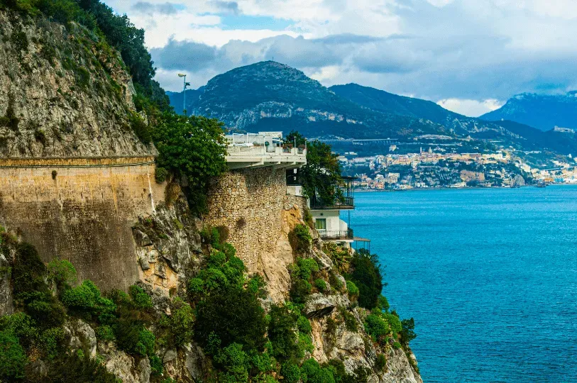 A winding coastal road and stone terraces built directly into the steep, rocky cliffs of the Amalfi Coast, with a distant coastal town visible across the bay.