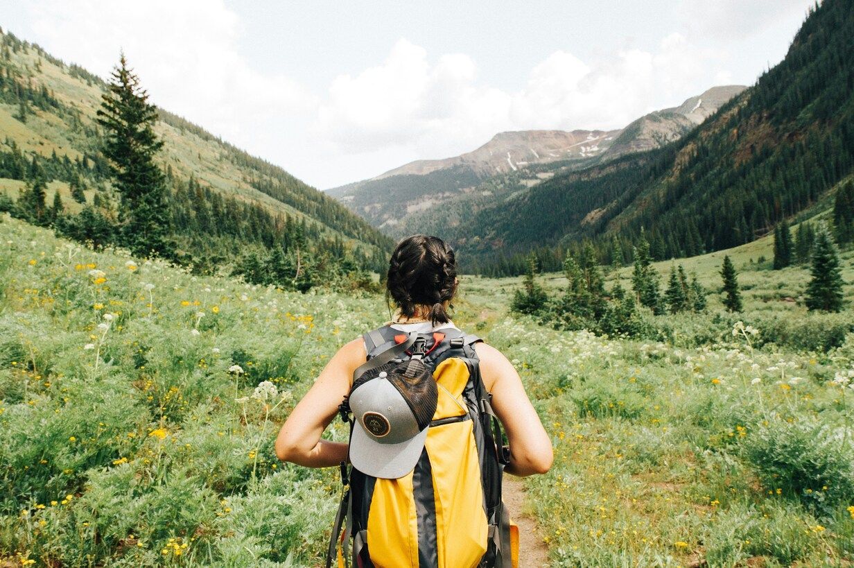 A woman wearing a bright yellow backpack hikes up a rocky mountain trail, surrounded by open sky and