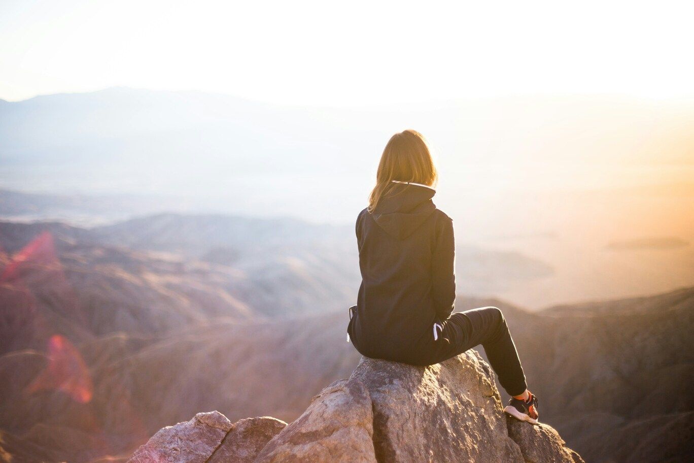 A woman sits quietly on a large rock overlooking a wide mountain landscape, with layered peaks and soft light in the distance, creating a peaceful and reflective travel scene.