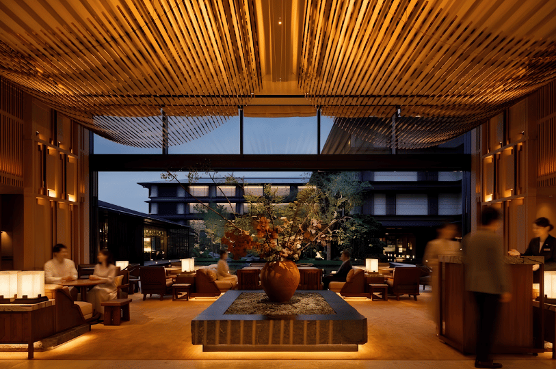 A spacious and sophisticated hotel lobby with a dramatic, symmetrical design featuring a ceiling of undulating wooden louvers. A grand central ikebana arrangement in a massive vase serves as a focal point. Seated guests are warmly lit, and large glass windows offer a view of a terraced building and a reflection pool at night.