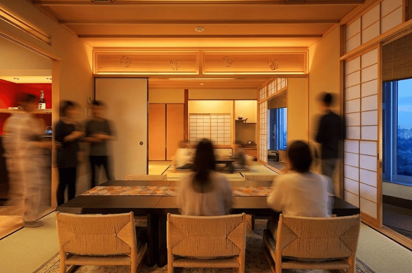 A high-end interior of a spacious traditional Japanese dining room featuring tatami mats, light wood construction, neat low tables, and warm cove lighting. A row of shoji paper screens along the right wall looks out onto a deep blue twilight sky, with blurred figures suggesting bustling guest activity.