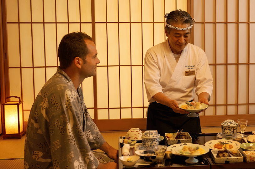 A male Japanese chef in a white jacket and patterned headband kneels and respectfully presents a beautifully plated dish with both hands to a seated guest in a grey patterned yukata. The setting is an intimate traditional Japanese room with tatami mats, shoji screens, and a small Japanese lantern.