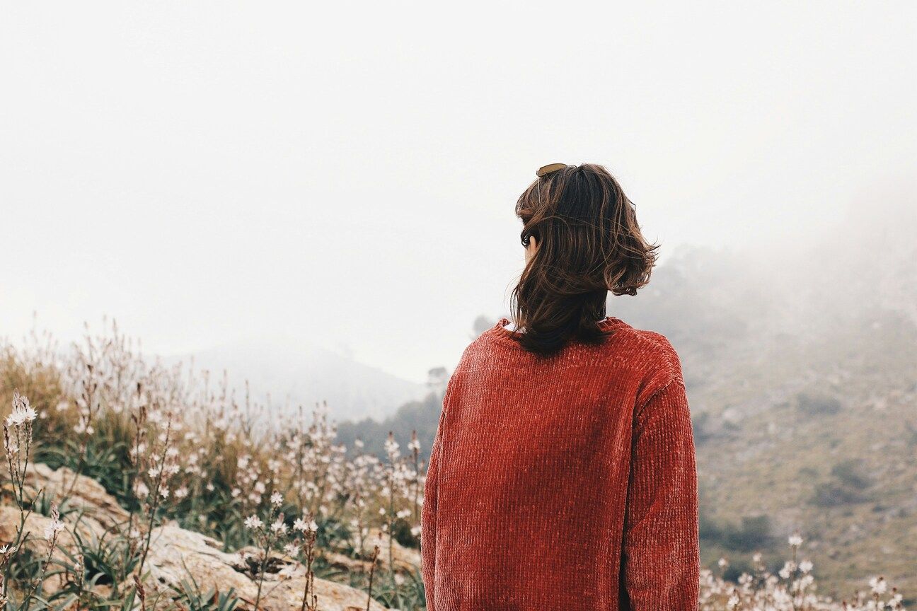 A woman wearing a backpack looks toward the mountain landscape, suggesting a moment of travel and reflection.