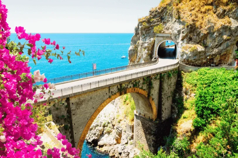 Vibrant pink bougainvillea flowers frame a picturesque arched stone bridge on a coastal road. The ro