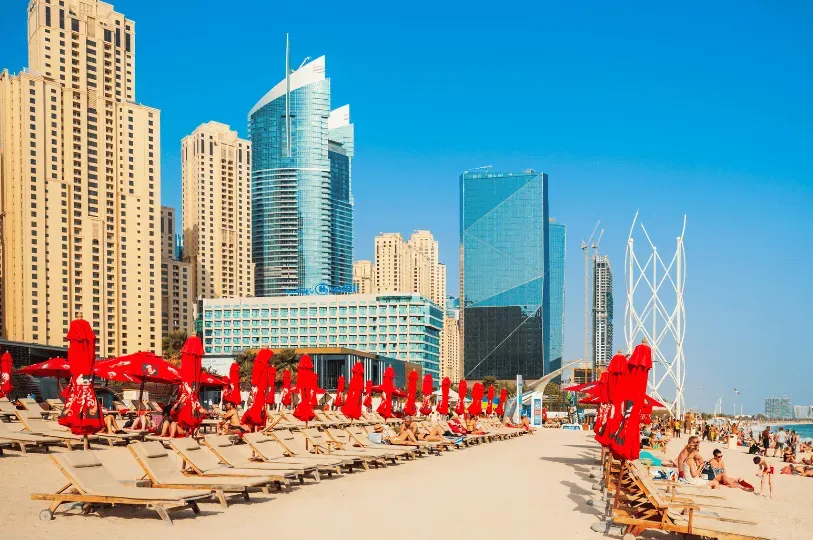  Rows of wooden lounge chairs and bright red umbrellas line a bustling beach in Dubai, contrasting w