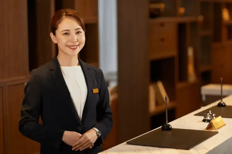 A welcoming, smiling female hotel receptionist in a professional black suit standing ready behind a modern marble front desk.