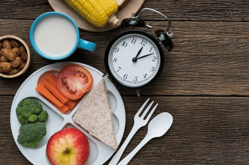 A top-down view of a healthy meal plate with a sandwich, broccoli, carrots, and an apple next to an analog alarm clock set to 1:10. This visual emphasizes the importance of timing meals to manage circadian rhythms.