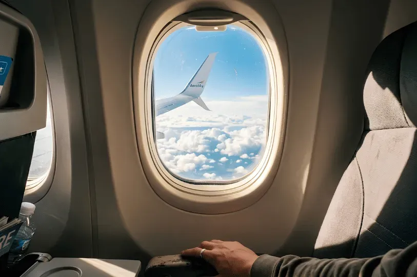 A view through an airplane window showing the wing of the plane and a thick layer of white clouds under a bright blue sky. The sunlight streaming into the cabin illustrates the exposure to natural light while traveling.