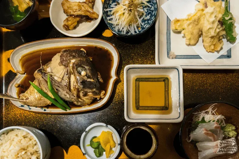 An overhead view of a diverse kaiseki spread on a black and gold lacquered tray, showcasing seasonal ingredients including a braised fish head, delicate sashimi, crisp tempura, and pickled vegetables.