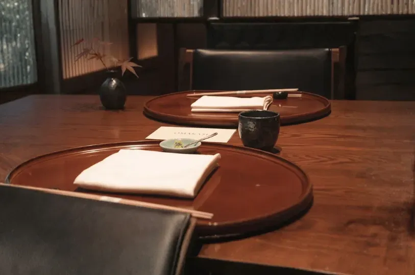 A minimalist wooden table setting for a kaiseki dining experience, featuring round lacquered trays, folded white linen napkins, chopsticks, and a simple vase with a red maple branch reflecting the changing seasons.