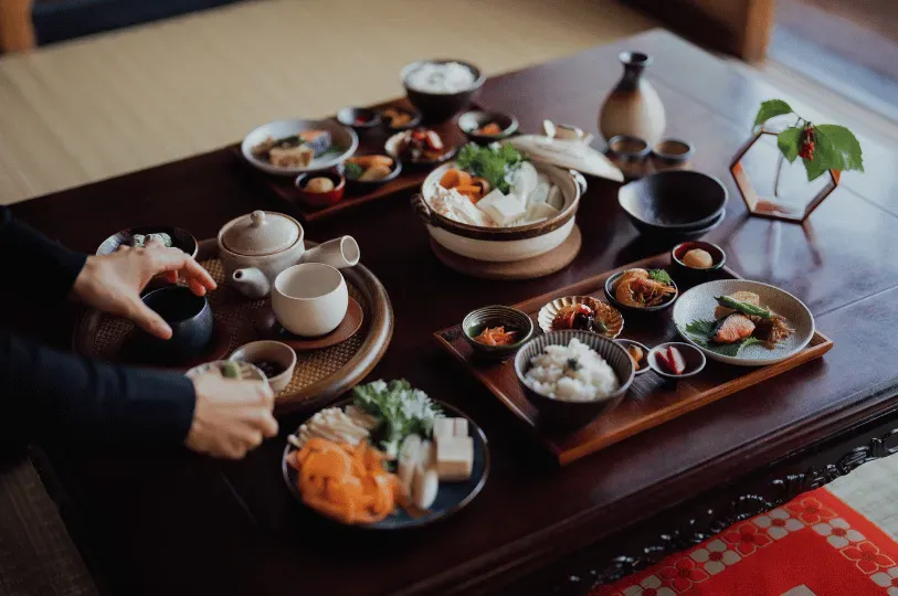 Hands carefully arranging a multi-course kaiseki meal on a low wooden table, highlighting a hot pot, fresh vegetables, grilled fish, and rice served in artisanal ceramics to emphasize seasonal harmony.