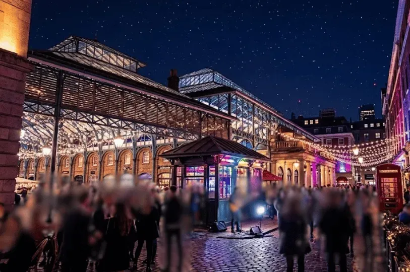 A bustling courtyard under a historic iron and glass market roof in London at night, adorned with warm string lights. A large, blurred crowd of people fills the cobblestone plaza beneath a starry night sky.