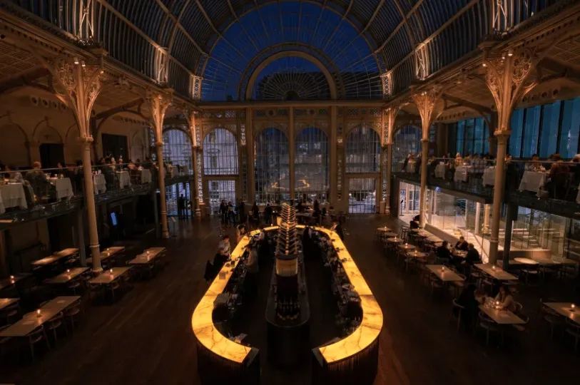 An elegant, multi-level bar and dining space inside a grand conservatory with high arched glass ceilings and ornate ironwork. A long, illuminated yellow bar anchors the center, with upscale patrons mingling and dining under warm ambient lighting.