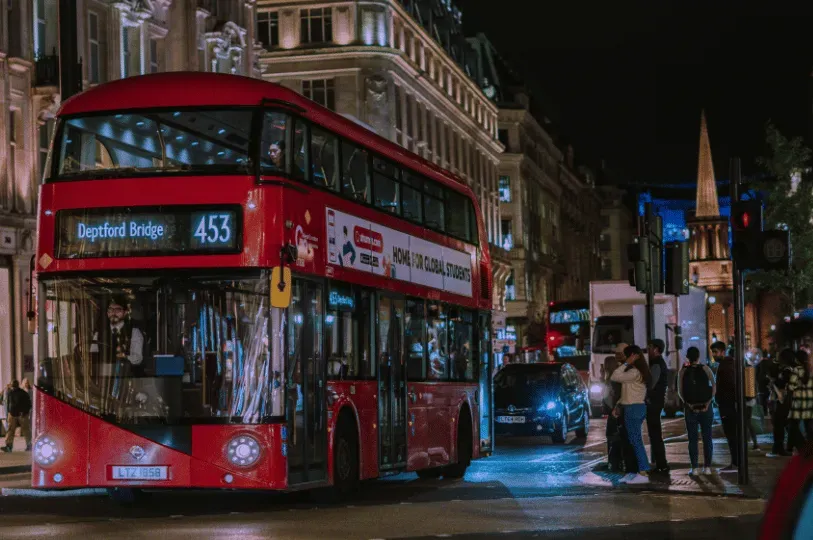 A classic red double-decker London bus, displaying route 453 to Deptford Bridge, traveling down a busy city street at night. Pedestrians stand on the brightly lit sidewalk near a crosswalk.