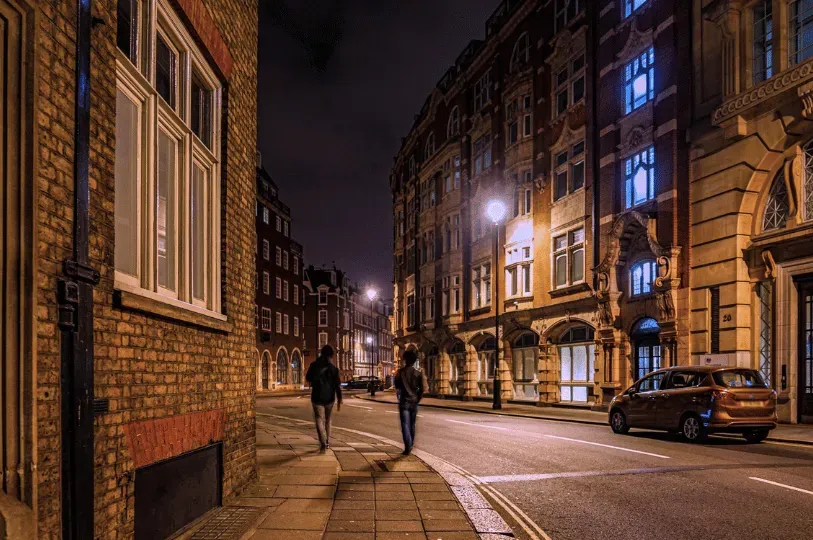 Two silhouetted figures walking away down a quiet, historic brick-lined street in London at night. The curved street and buildings are illuminated by the warm glow of classic streetlamps.