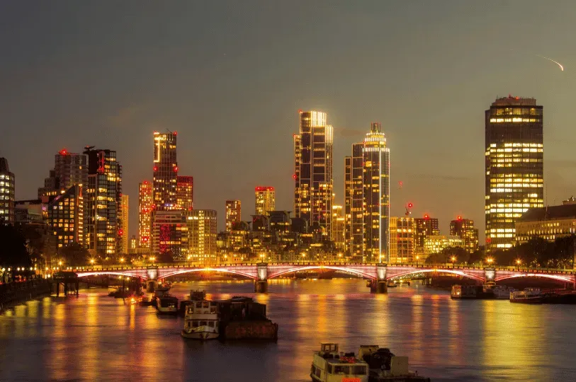 A glowing nighttime skyline of London across the River Thames. A bridge brightly illuminated with red lights spans the calm, reflective water, with boats moored in the foreground and modern skyscrapers towering in the background against a twilight sky.