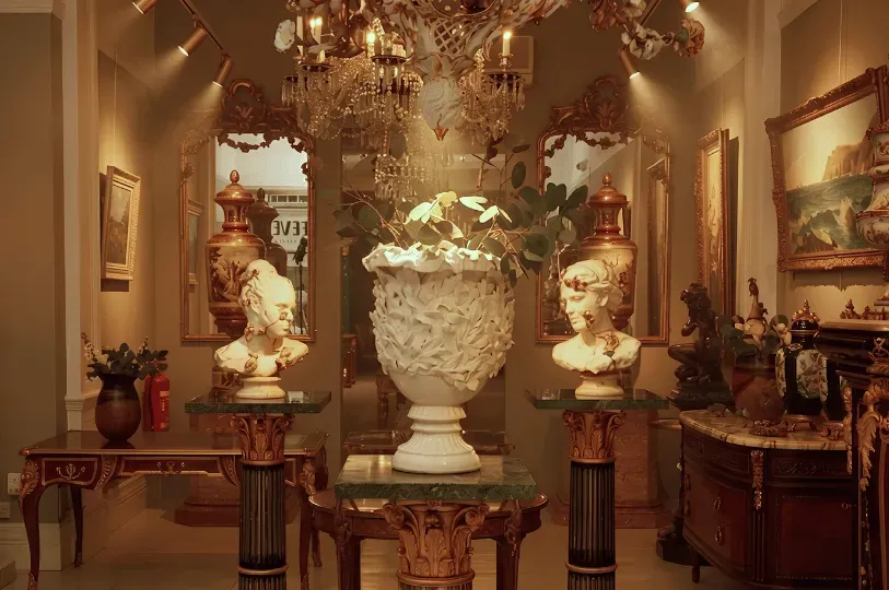An opulent, warmly lit private gallery interior featuring classical marble busts on fluted columns, a large ornate chandelier, gilded mirrors, and fine antique mahogany furniture.