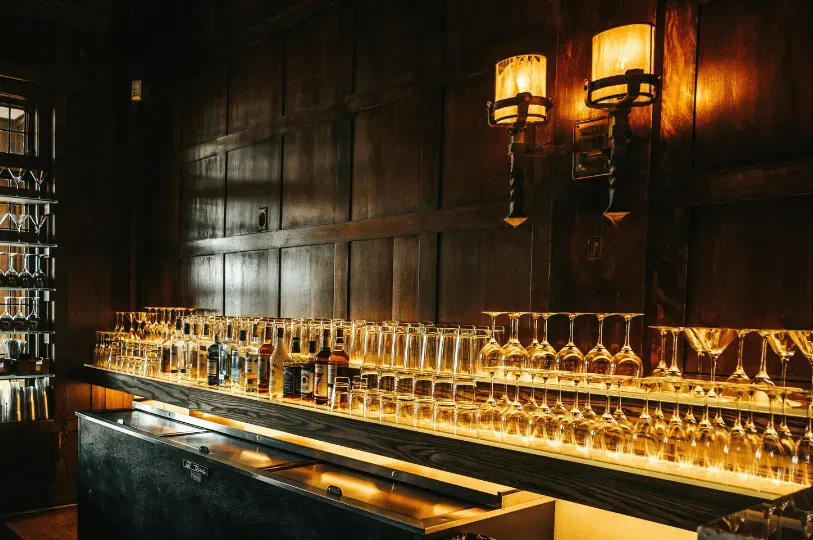  Warm, vintage wall sconces illuminate neat rows of clean glassware resting on glowing shelves against dark wood paneling inside a moody speakeasy.