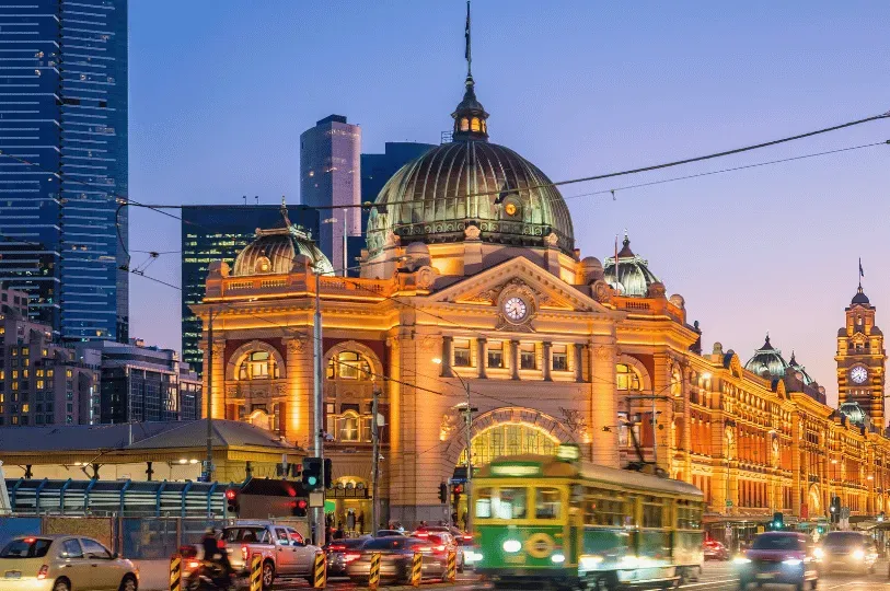The iconic dome and facade of Melbourne's Flinders Street Railway Station brightly illuminated at dusk, with a classic green and yellow tram passing through city traffic in the foreground.
