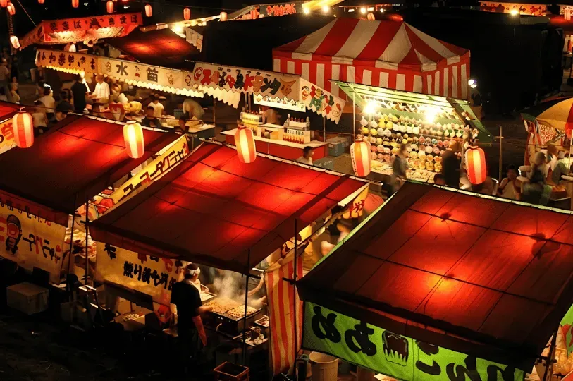 Vibrant night scene at a Japanese summer matsuri, featuring crowded, brightly lit food stalls with red awnings and glowing paper lanterns, with a vendor cooking street food in the foreground.