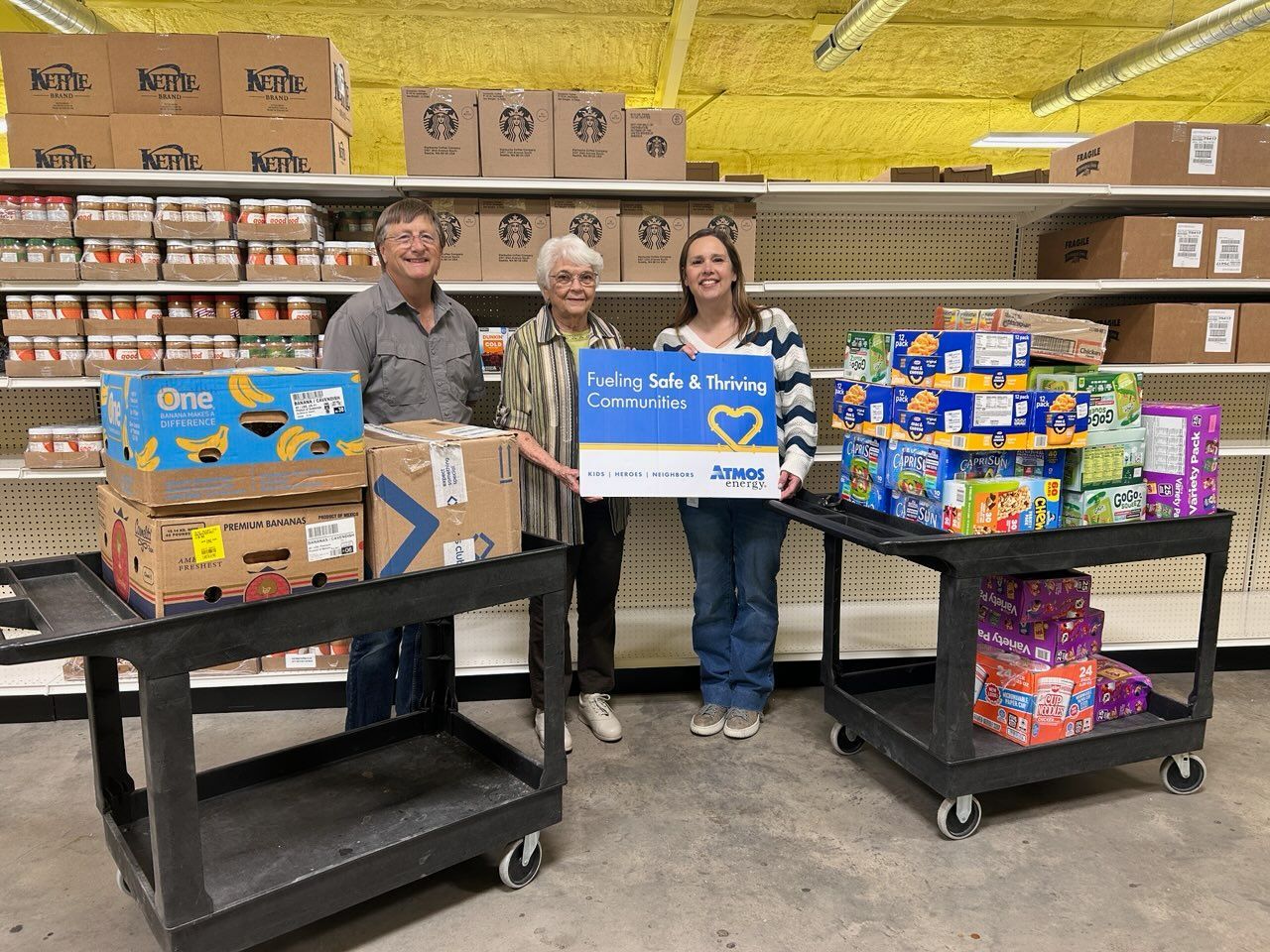 Three people stand in a pantry holding boxes for a food drive, with shelves of supplies in the background.