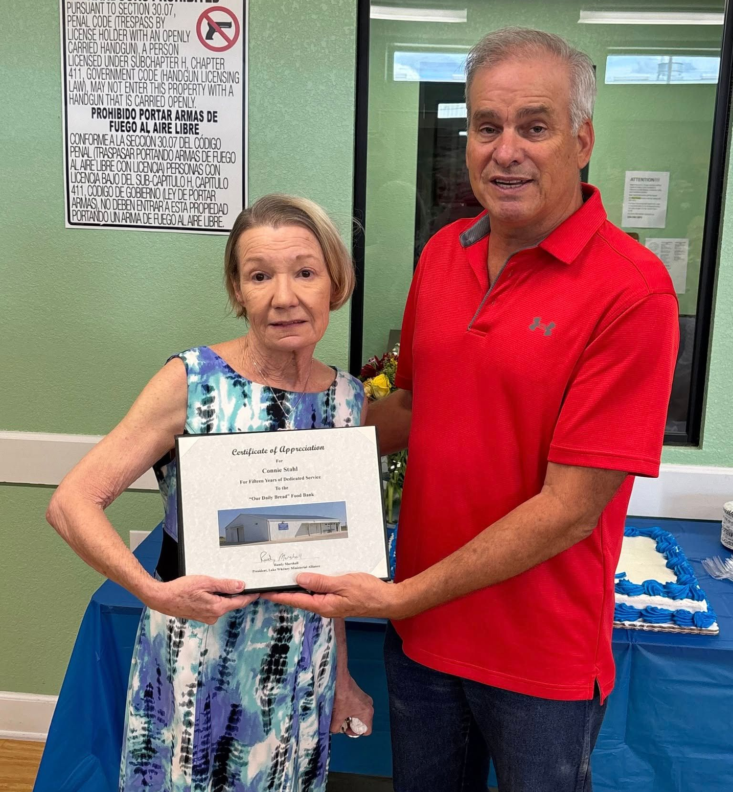 Two people stand side-by-side, holding a framed certificate in front of a blue-covered table in an indoor setting.