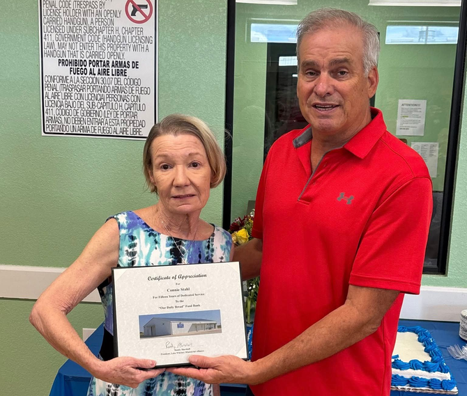 Two people stand side-by-side, holding a framed certificate in front of a blue-covered table in an indoor setting.
