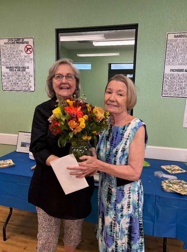 Two women smiling, one holding flowers and a card. Indoors, with a blue tablecloth and wall decor.