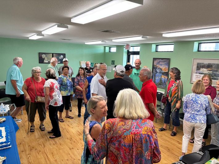 A group of people gathers in a room with light green walls and light wood floors, conversing at an indoor social event.