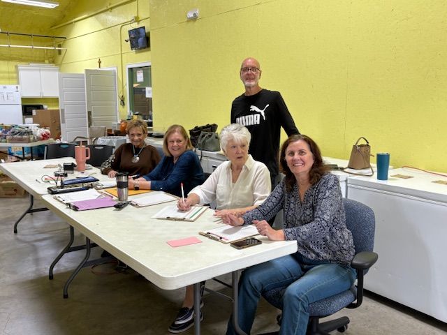 Five people seated around a table, one standing behind. People writing, light yellow wall.