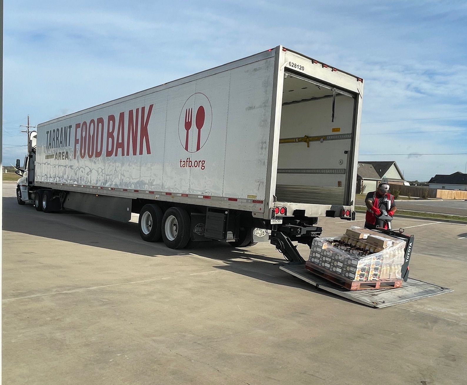 A person in a red jacket uses a pallet jack to unload a pallet of food from a white Tarrant Area Food Bank semi-truck.