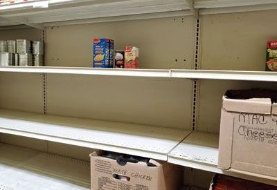 Empty store shelves with a few remaining food boxes and two cardboard shipping boxes on the bottom shelf.