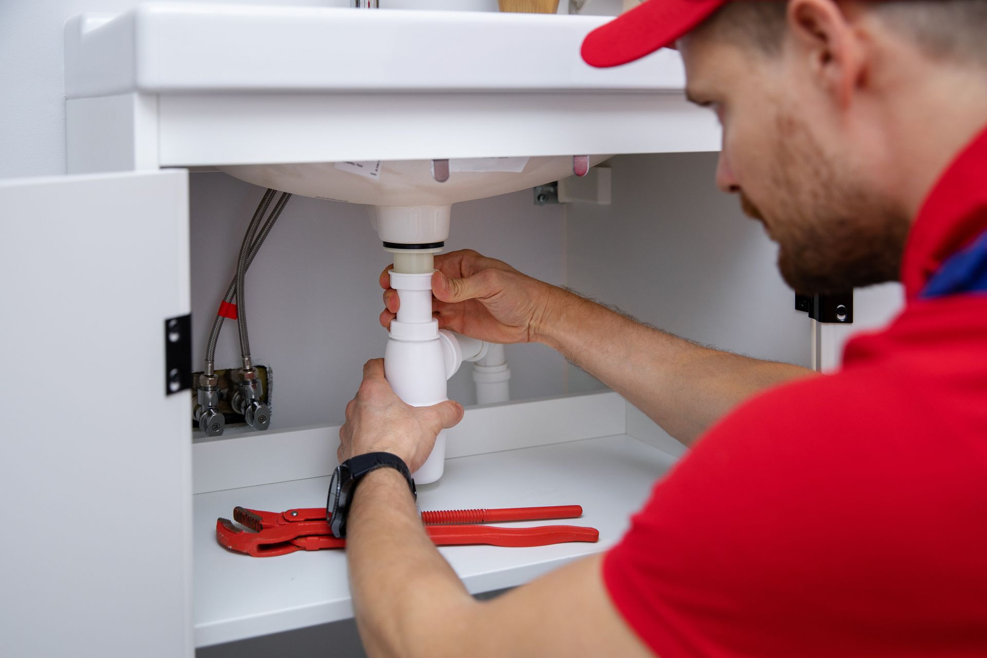 Male emergency residential plumber with red uniform installs white sink siphon in bathroom. Male emergency residential plumber with red uniform installs white sink siphon in bathroom.