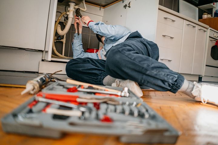 A plumber is kneeling on the floor fixing a sink in a kitchen.