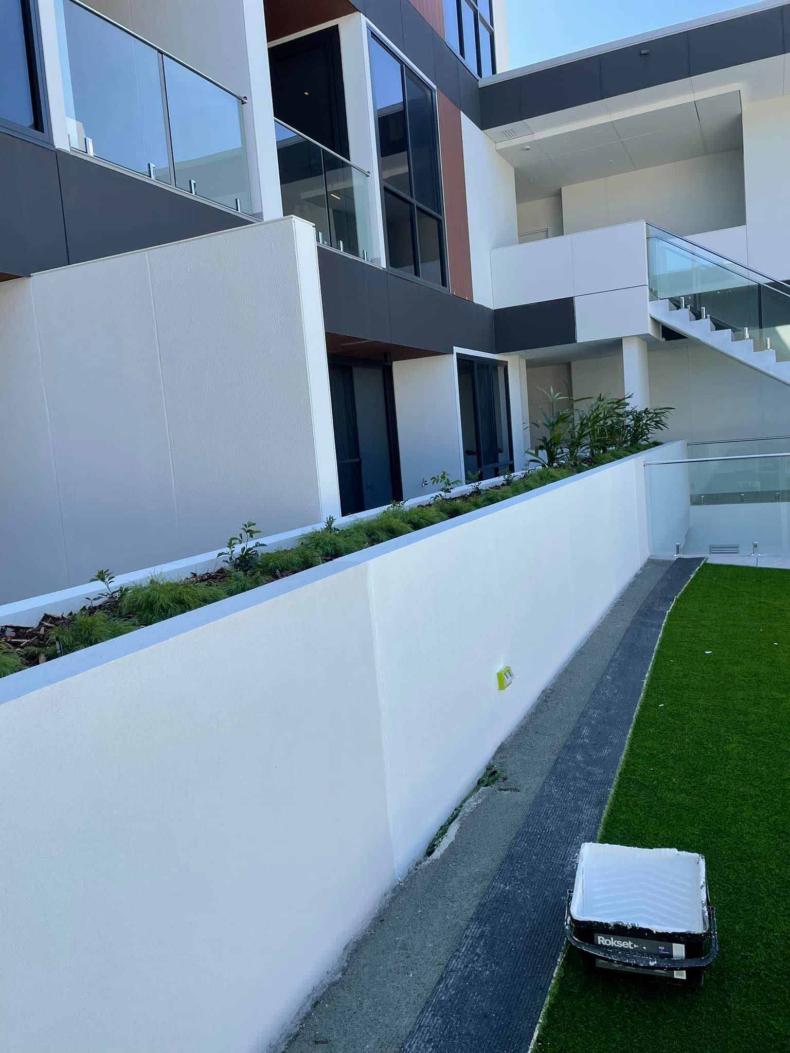 Modern building exterior with white walls, glass balconies, and a robot lawn mower on artificial grass.