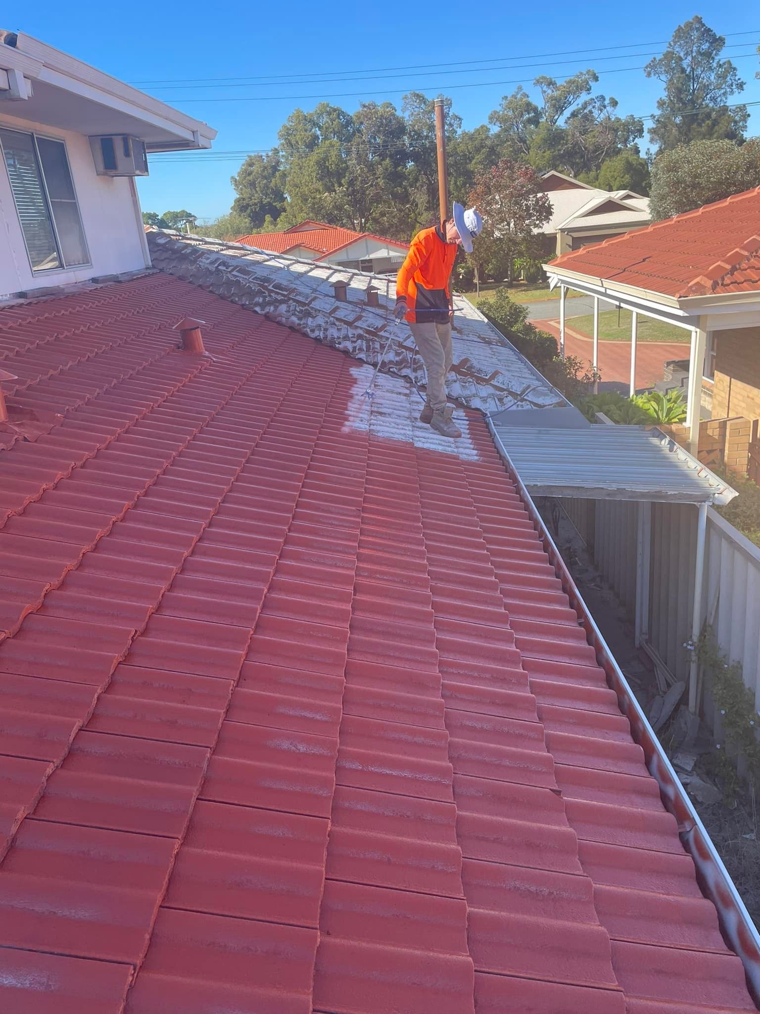 Person in orange vest on a red-tiled roof, applying white sealant near a gutter under a bright blue sky.