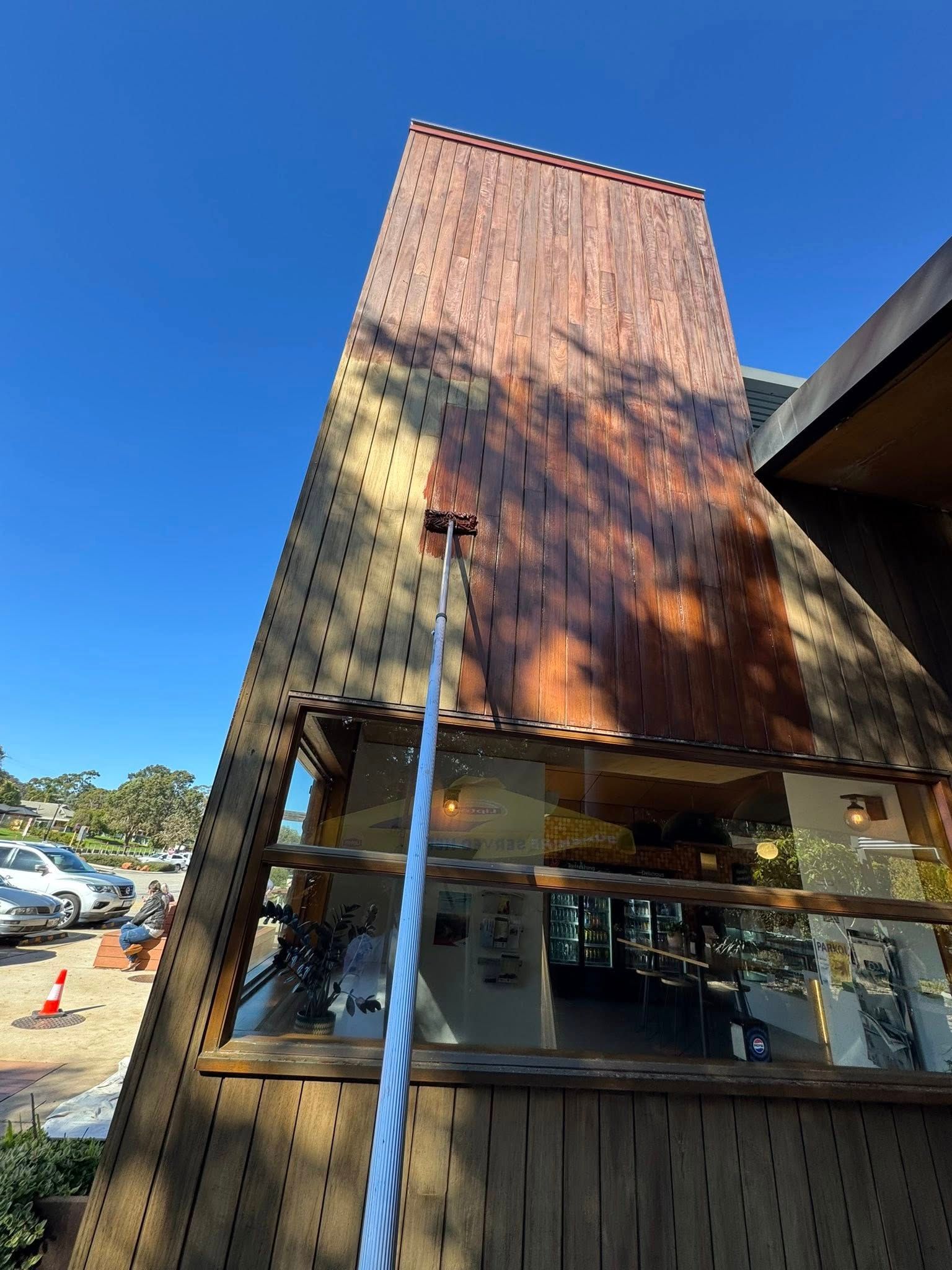 A person on a ladder washing the wood siding of a restaurant under a bright blue sky.