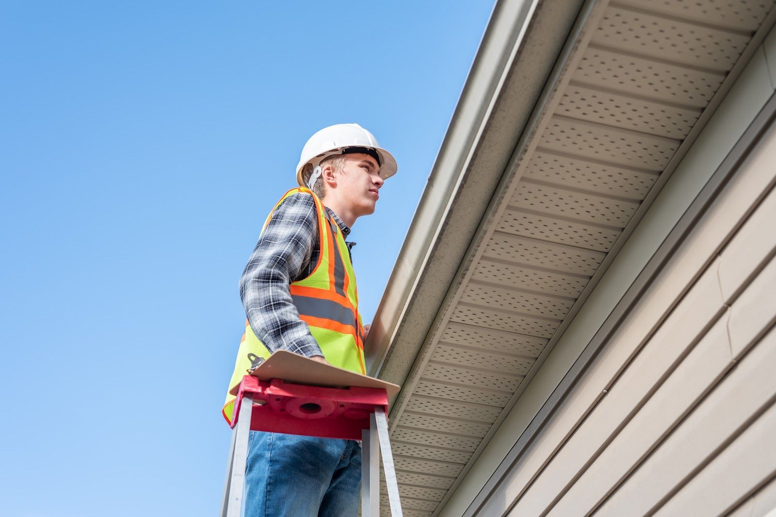 Man in hard hat and safety vest on a ladder inspecting a house's gutter under a blue sky.