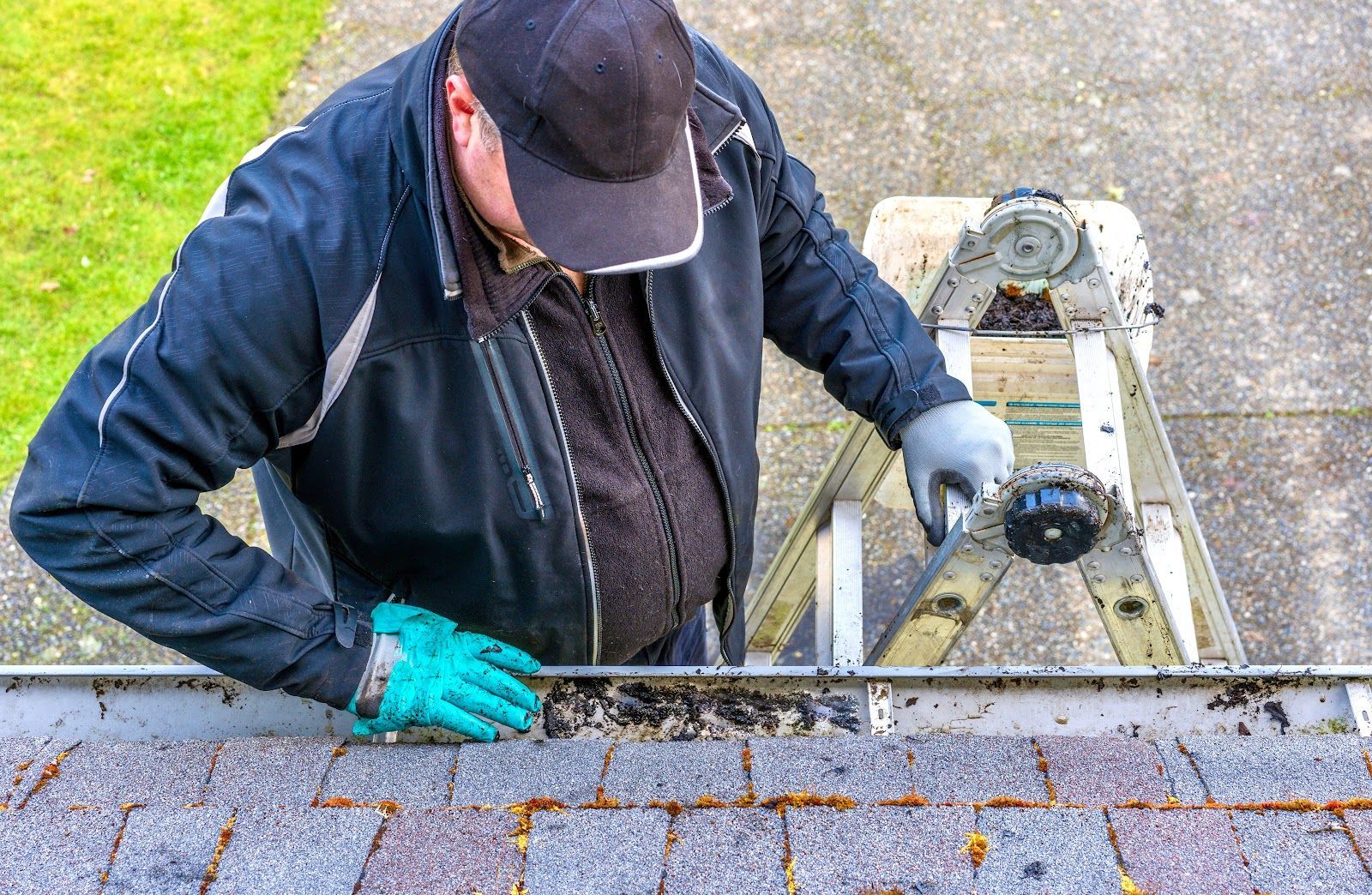 Person on a ladder cleaning a gutter; wearing gloves and a cap.