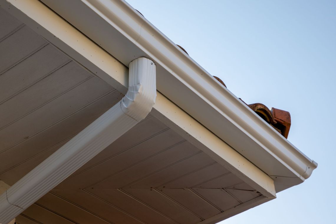 White gutter and downspout on a house roof corner against a blue sky.