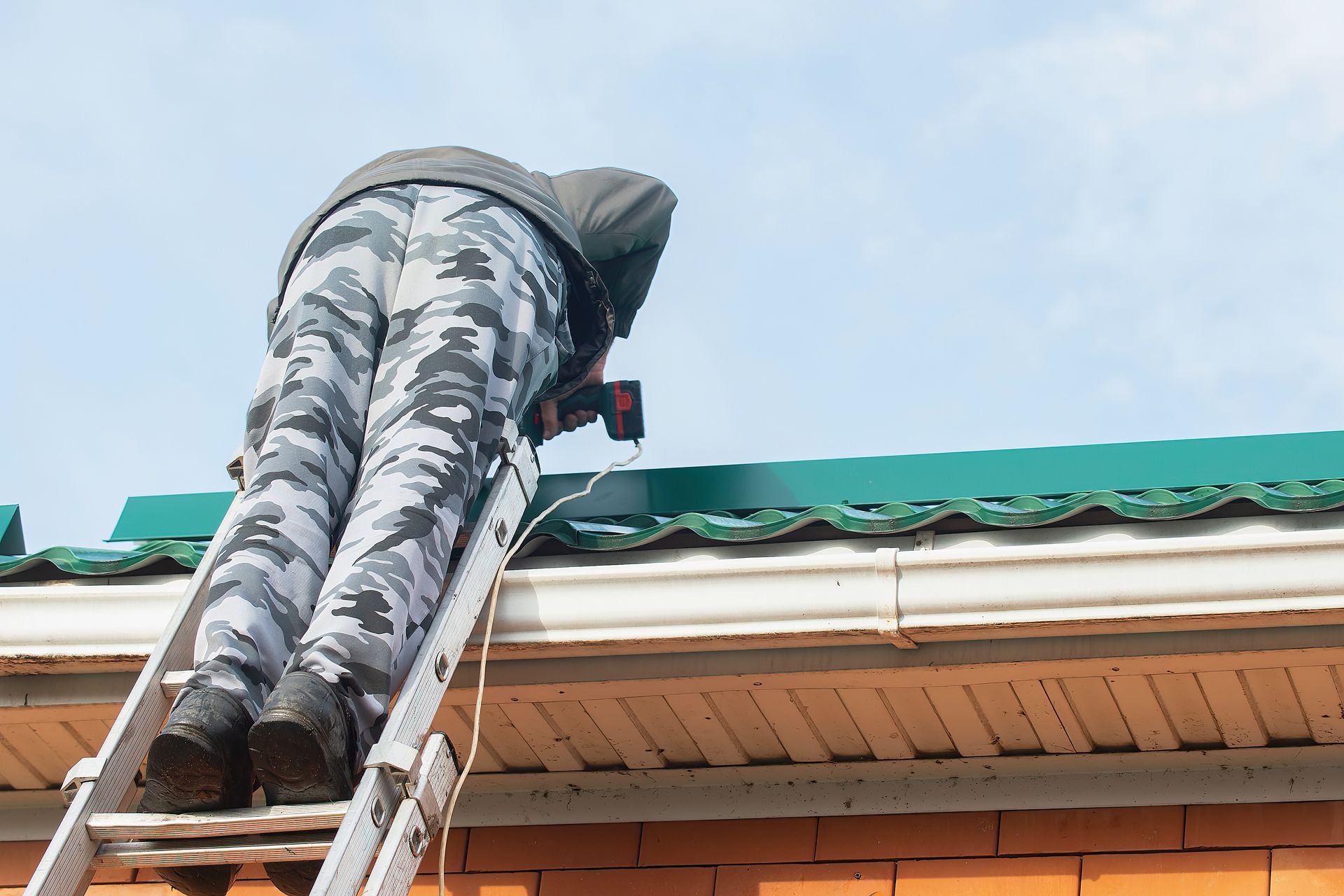 A person on a ladder working on a green roof with a power tool, blue sky.
