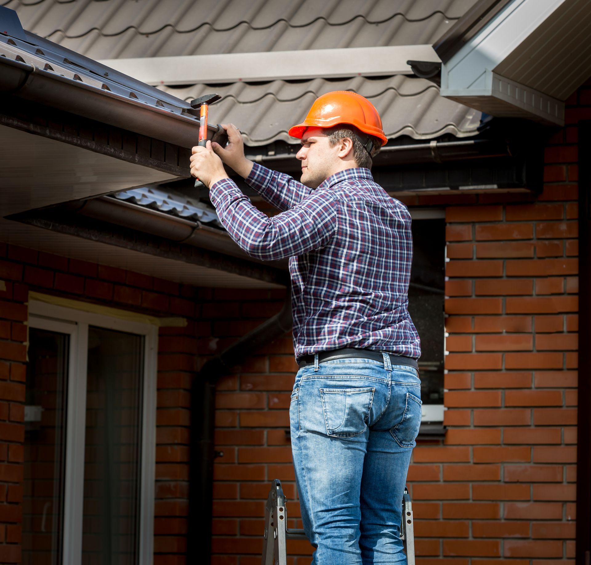 Man in hard hat on ladder hammering gutter. Red brick house, blue jeans, plaid shirt.