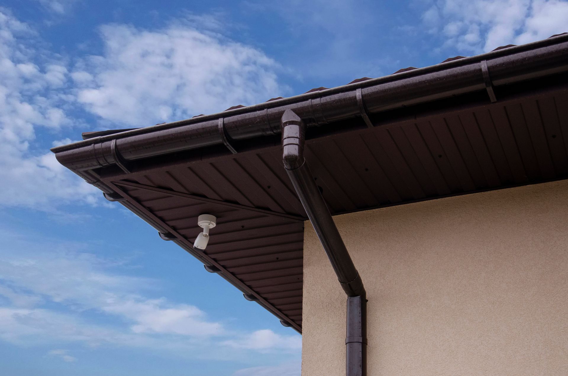 Brown roof with gutters and downspout on a light tan building against a blue sky.