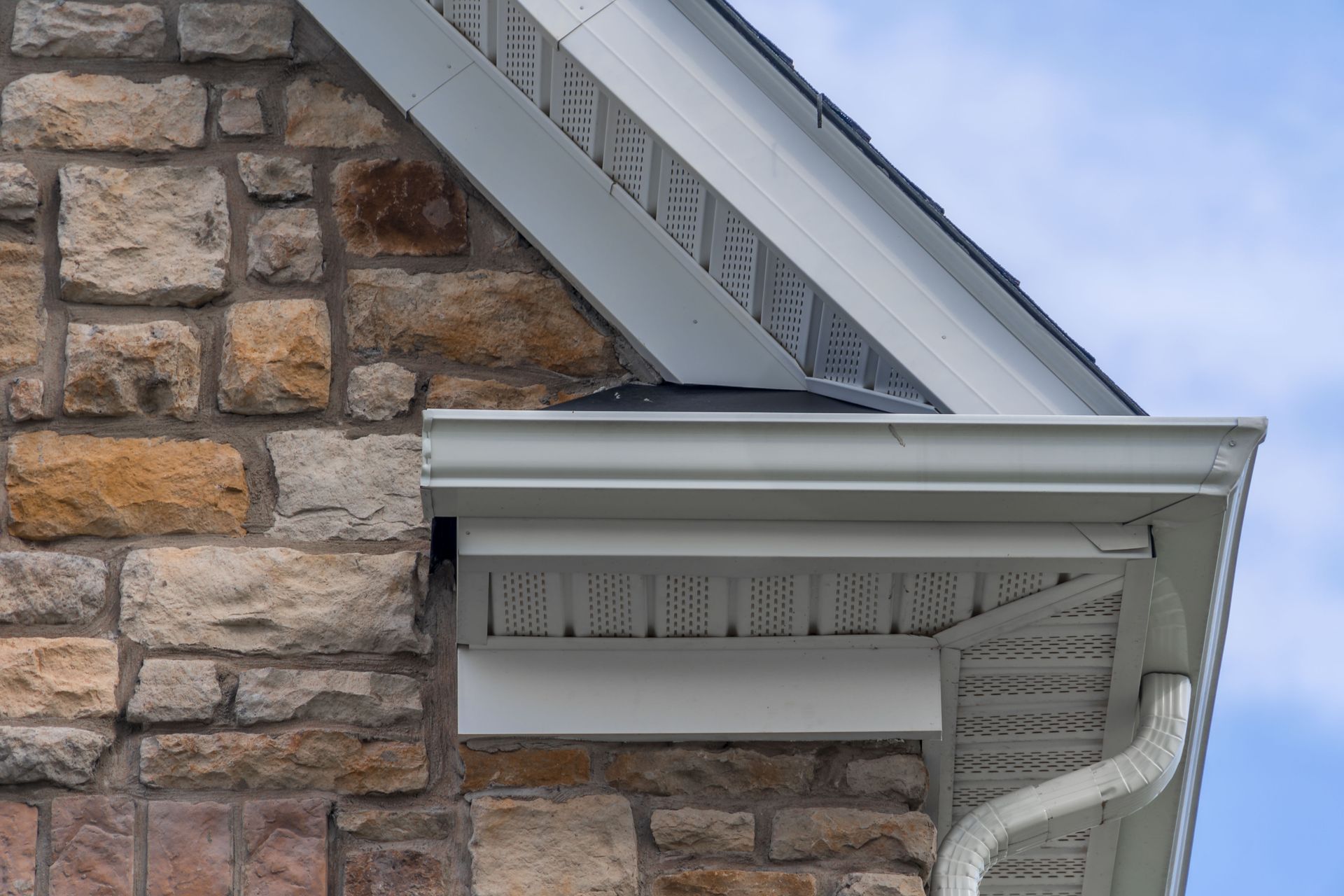 Stone brick wall with white gutters, soffit, and part of a roof against a blue sky.