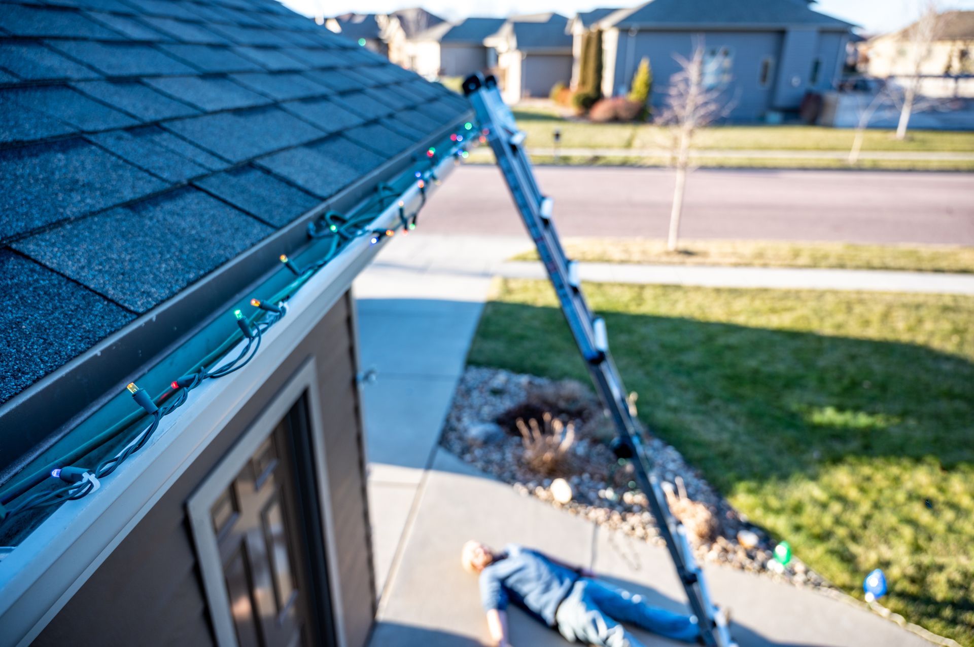 Person fallen on a sidewalk, ladder leaning on a house, Christmas lights on the roof.