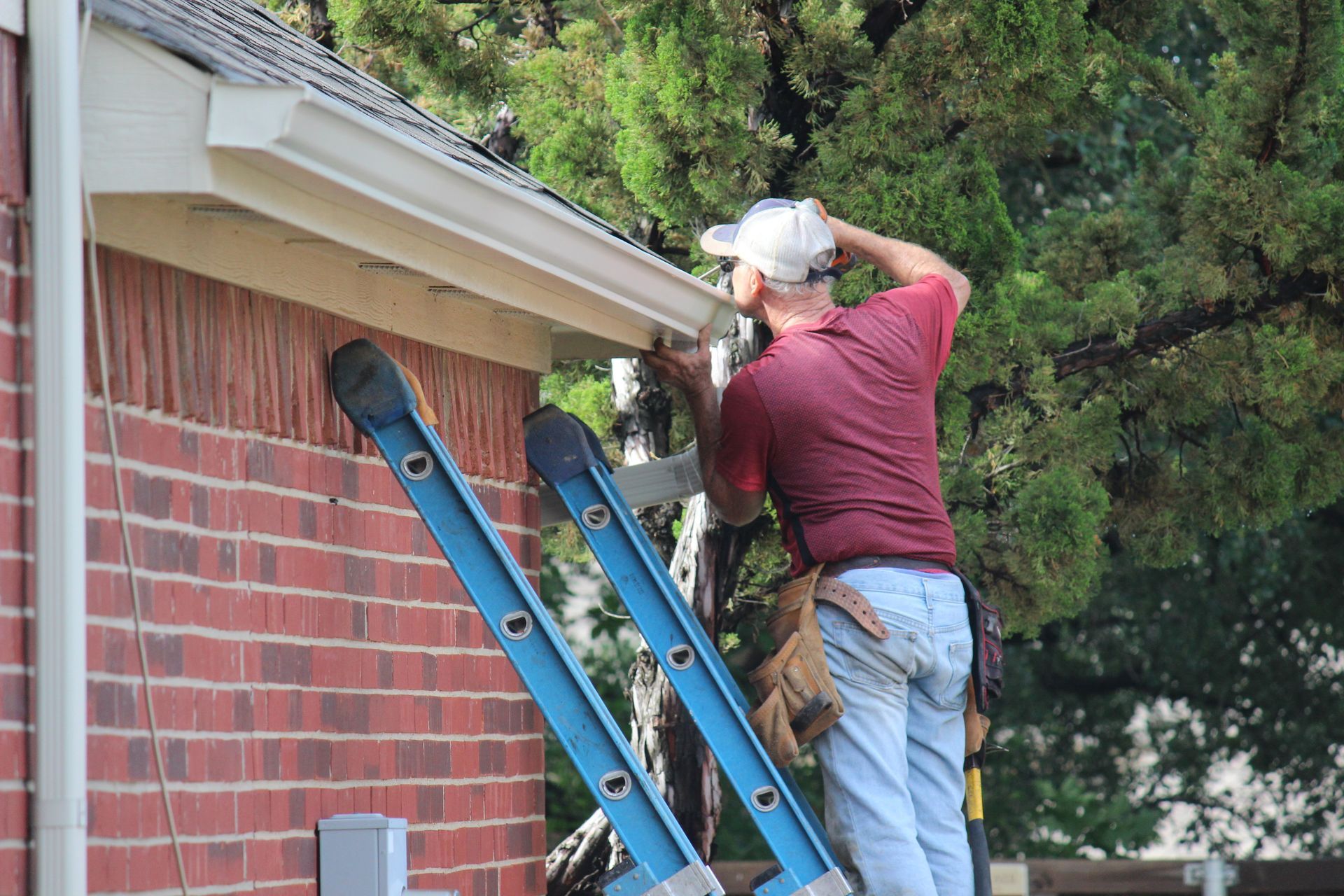 Man on ladder repairs a gutter on a brick house; sunny day.