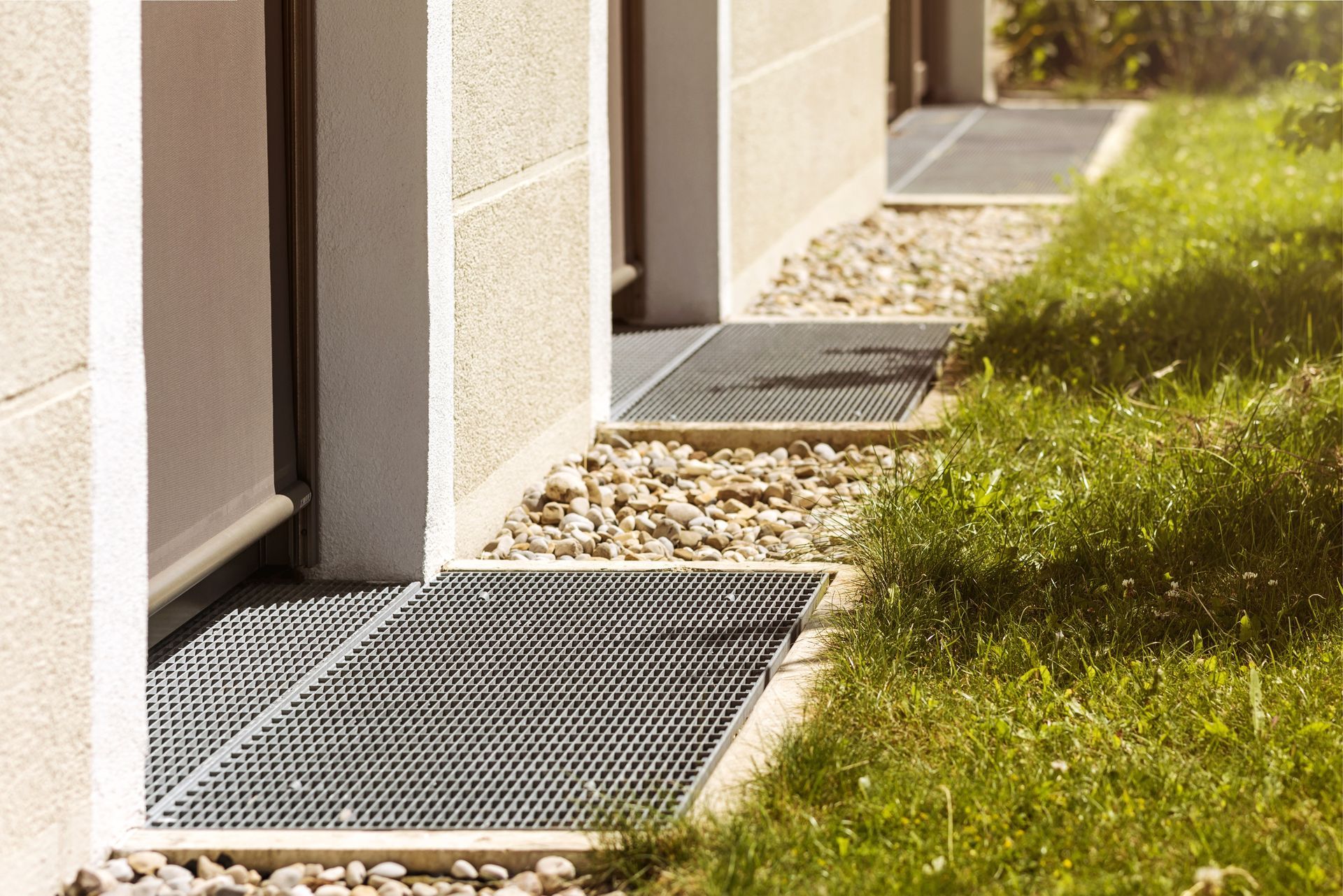 Exterior view of a building with grates in the ground near windows, set in gravel and grass.
