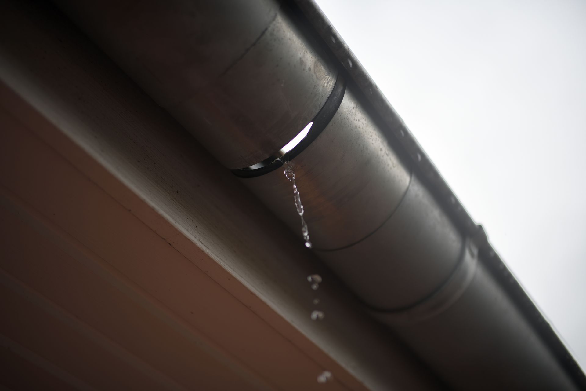 Leaking gray gutter with water droplets, mounted on a brown wall, overcast sky.